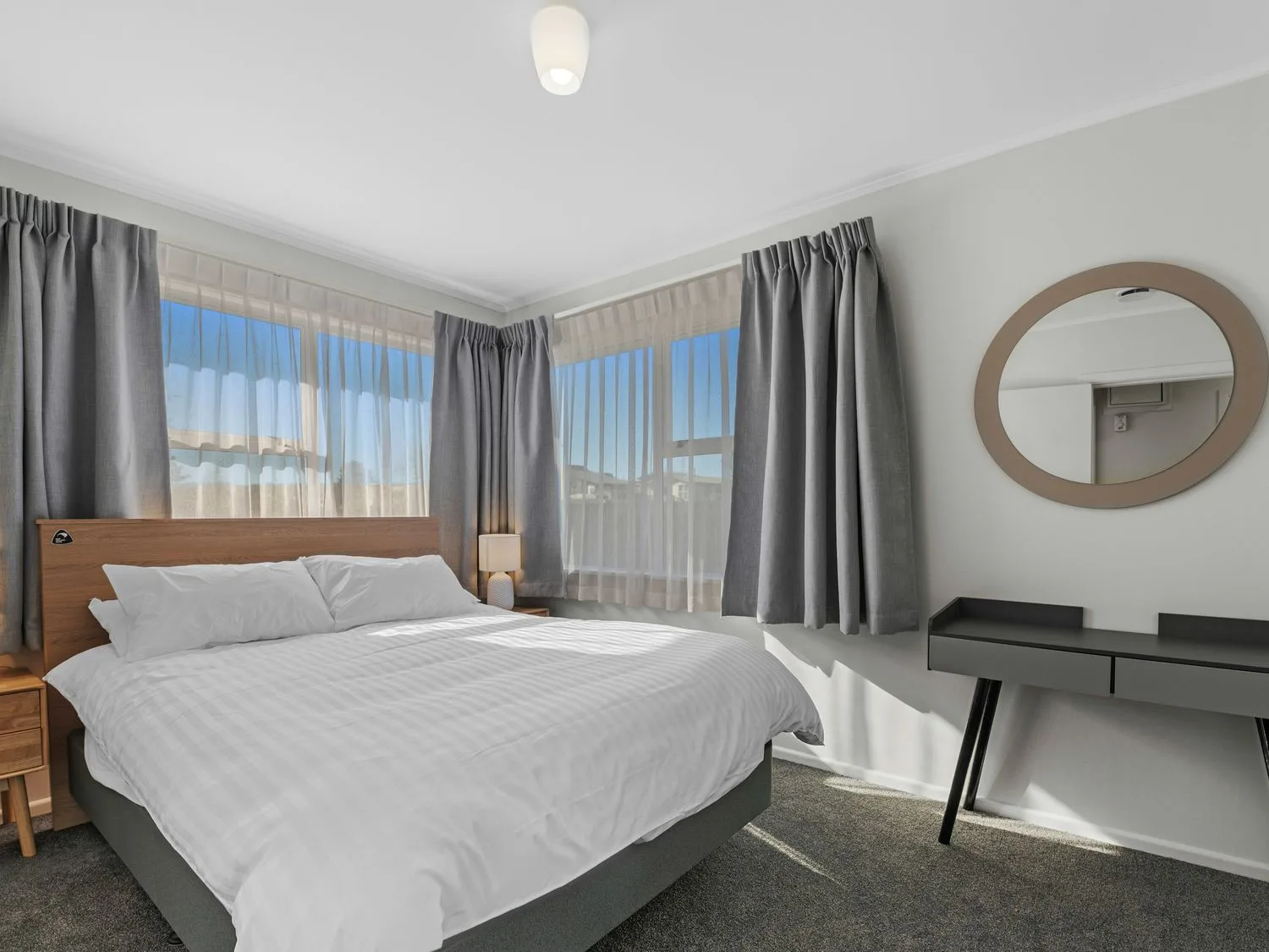 A bedroom with a bed a bedside table with lamp a desk with a round mirror and windows with curtains at Owhata Retreat - Rotorua Holiday Home in Rotorua