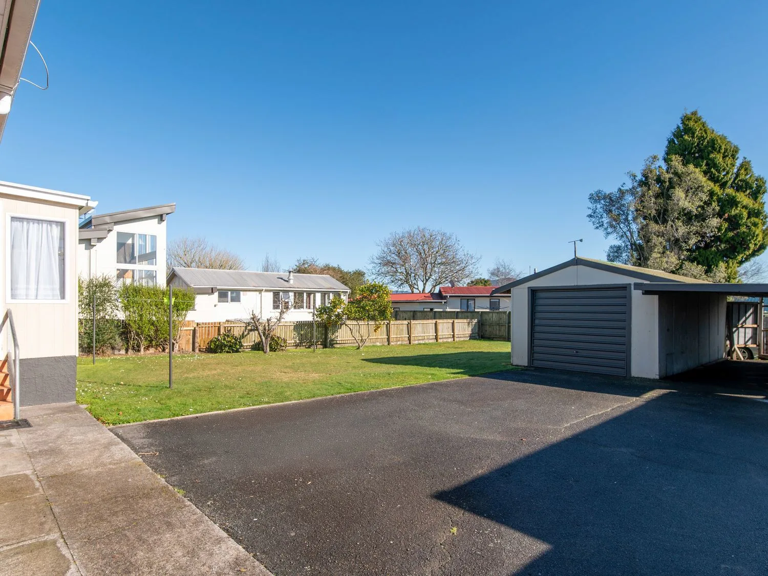 A driveway with a garage and adjacent lawn with trees and houses in the background at Owhata Retreat - Rotorua Holiday Home in Rotorua