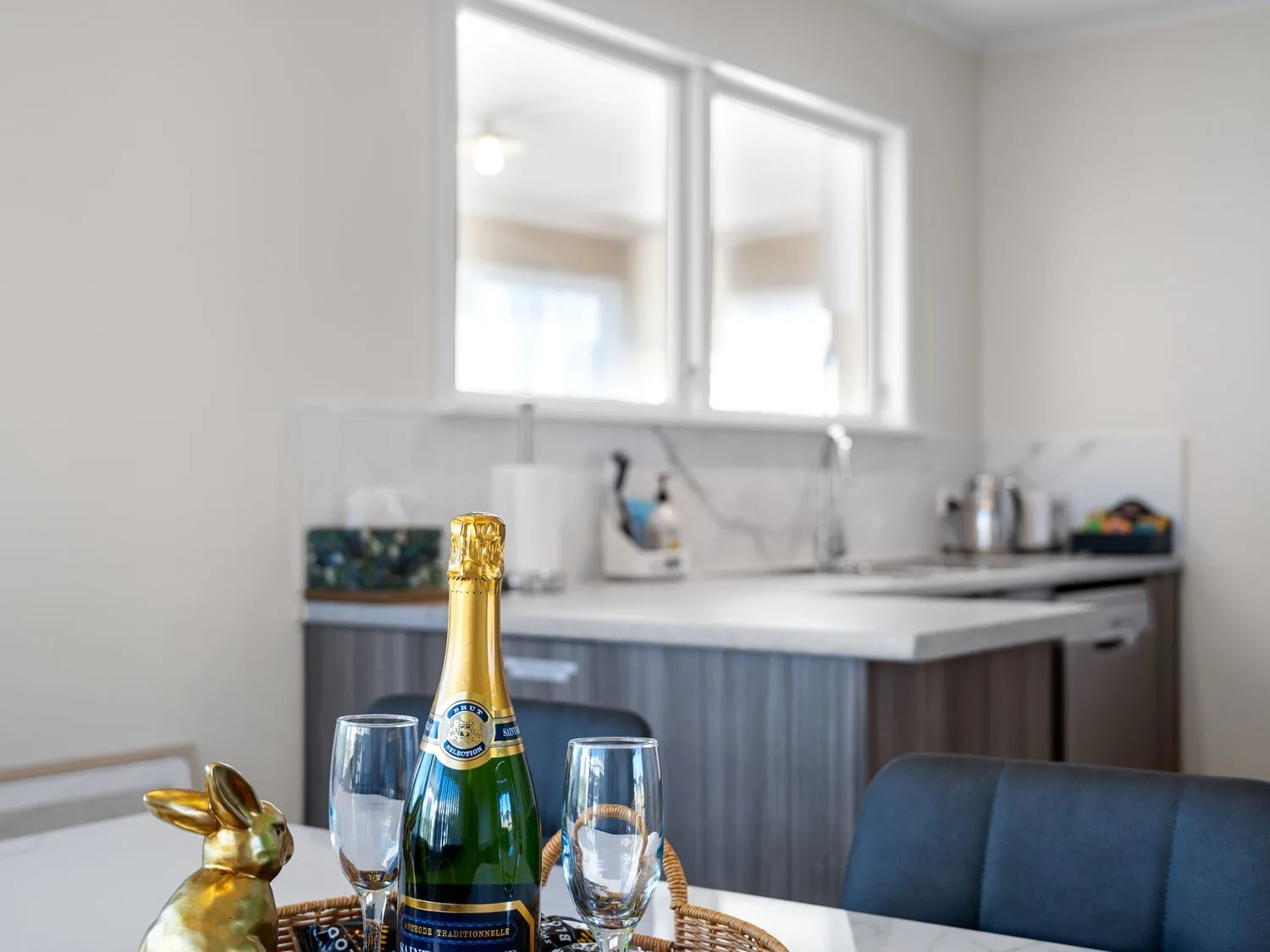 A kitchen with a countertop and sink with a bottle of champagne and two glasses on a table near a gold rabbit figurine at Owhata Retreat - Rotorua Holiday Home in Rotorua