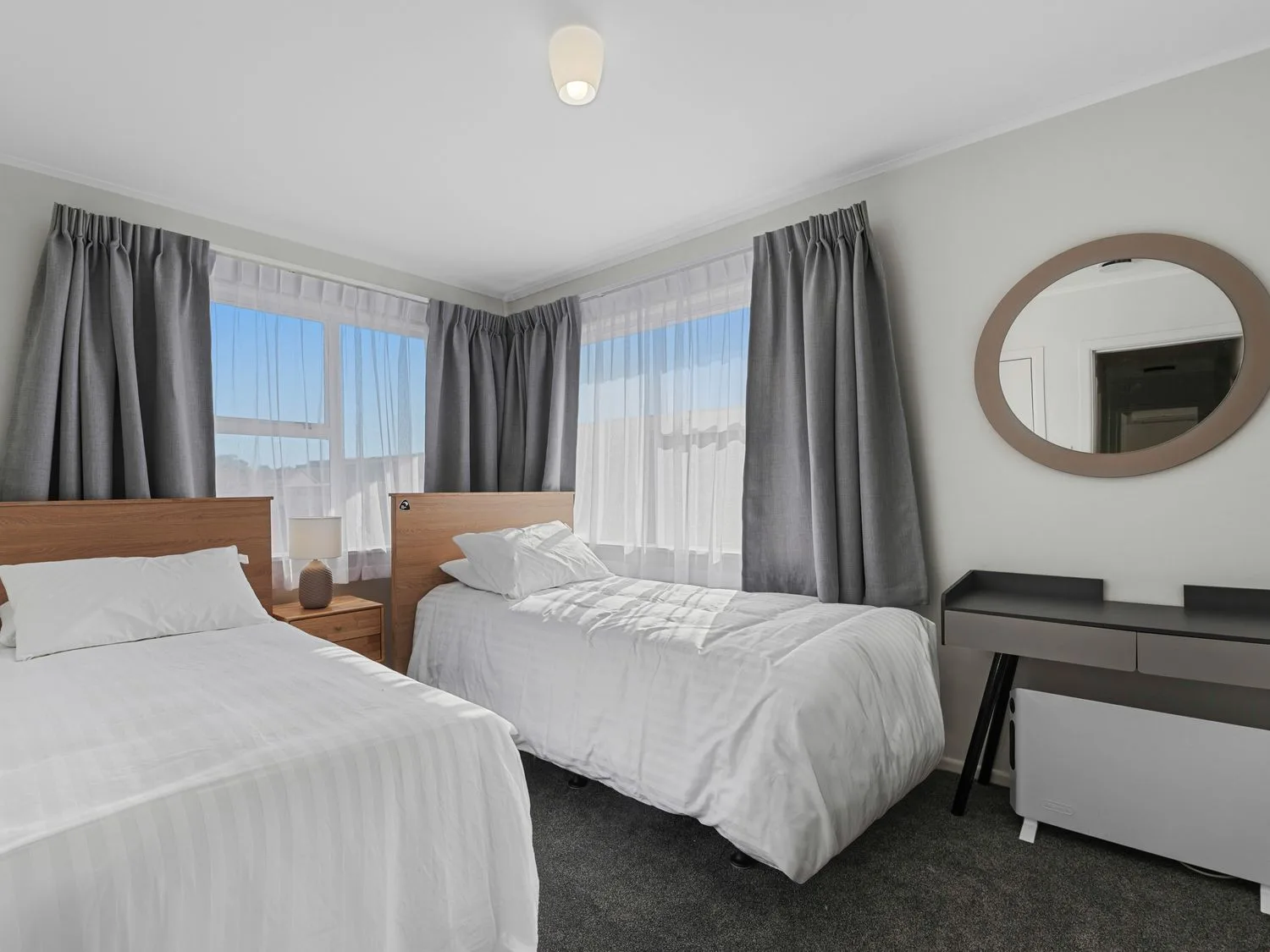 A bedroom with two single beds a bedside table with a lamp curtains a desk and a round mirror at Owhata Retreat - Rotorua Holiday Home in Rotorua