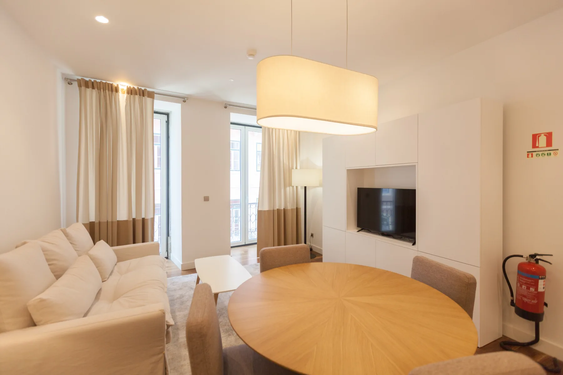 A living and dining area with seating, a television, and tall curtained windows.
