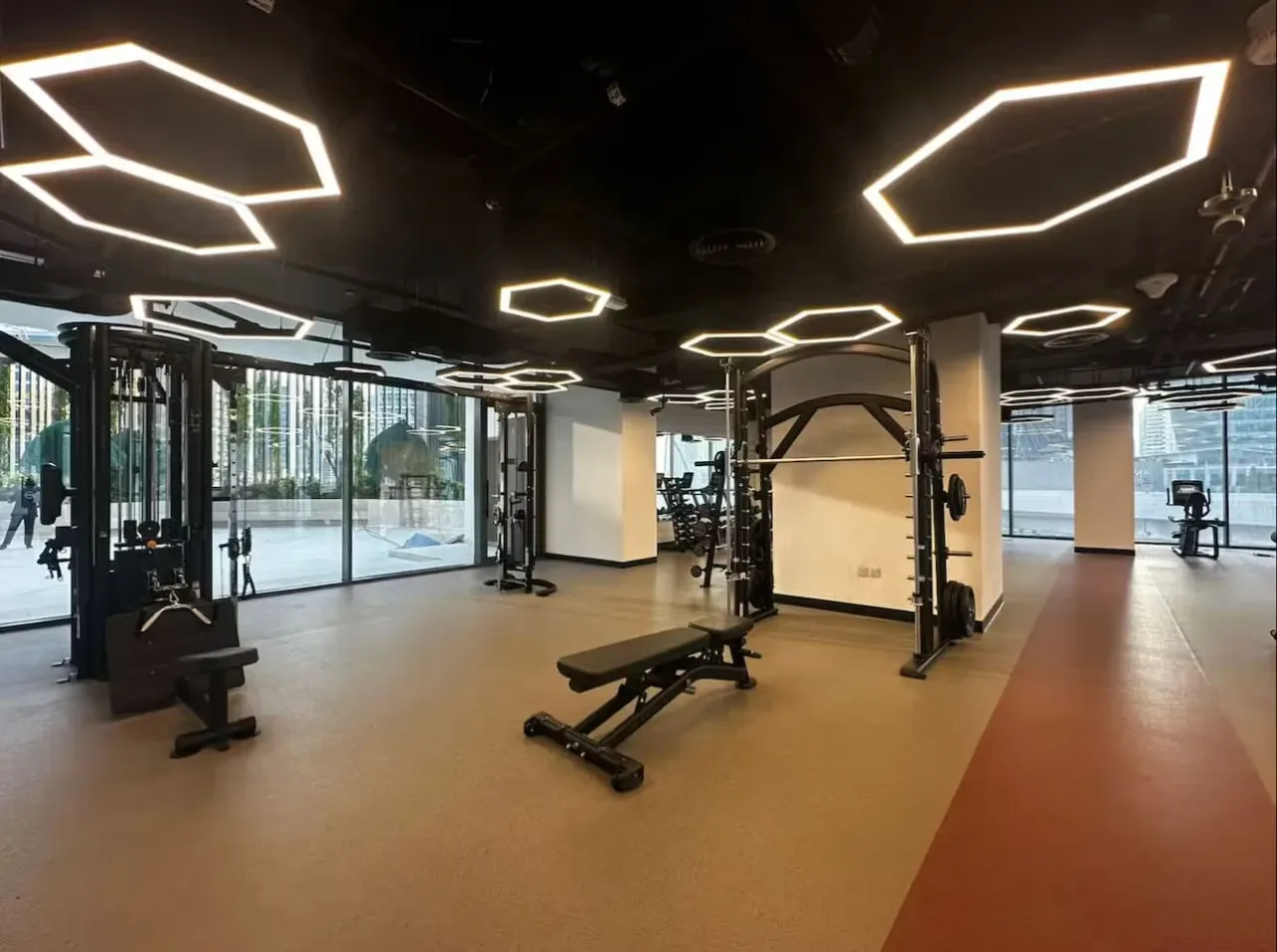 Un gimnasio con equipo de pesas y bancos junto a grandes ventanales.