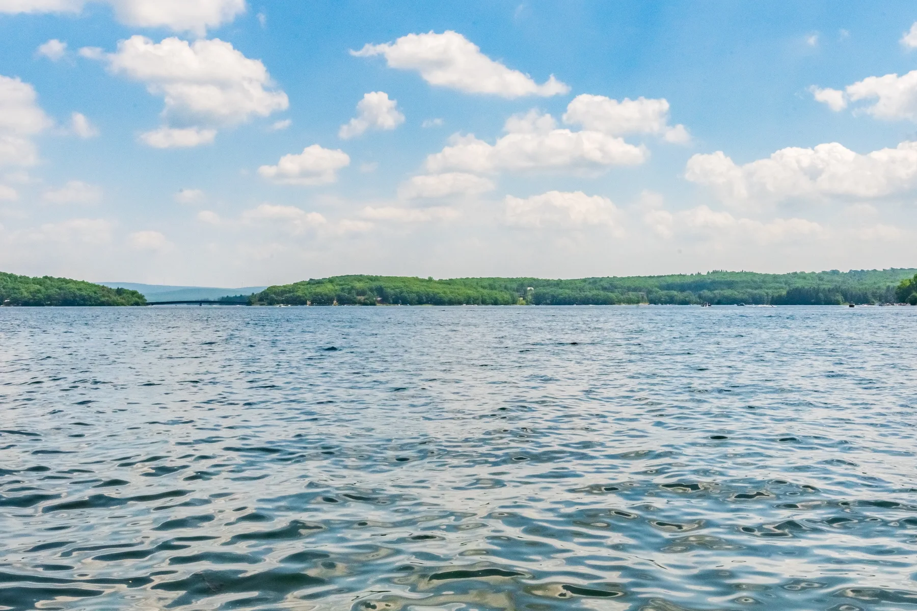 A broad lake stretches toward a green, tree-lined shore beneath a partly cloudy sky.