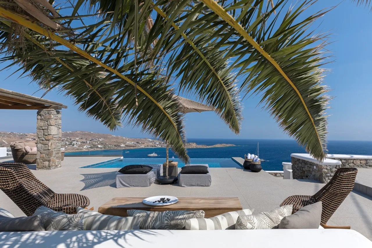 A seaside terrace overlooks the blue water, with outdoor seating beneath a palm tree.