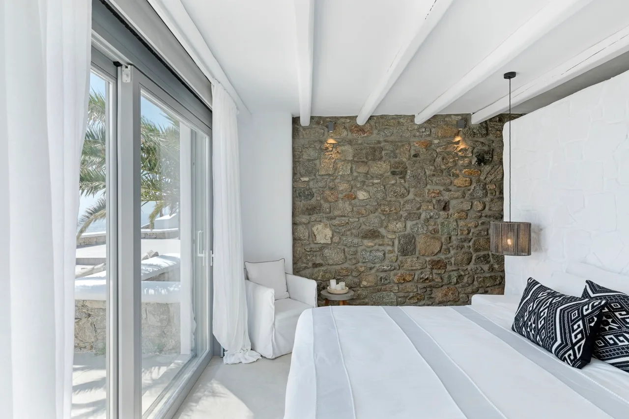 A bright bedroom with white bedding and a rustic stone accent wall.