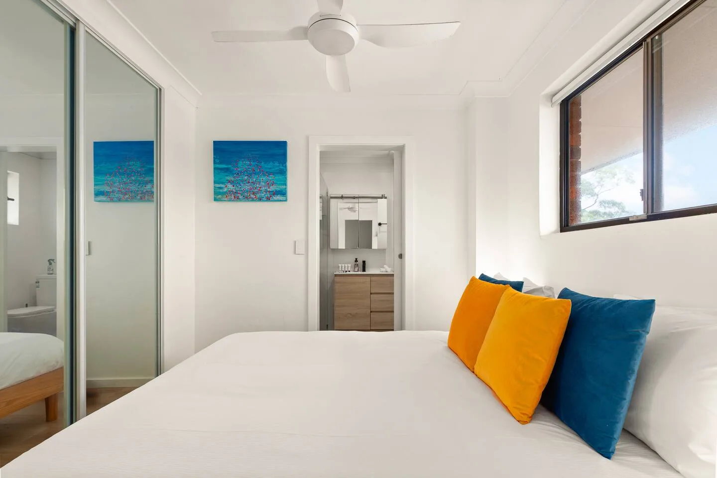 A bright bedroom with a white bed, colorful pillows, and a ceiling fan.