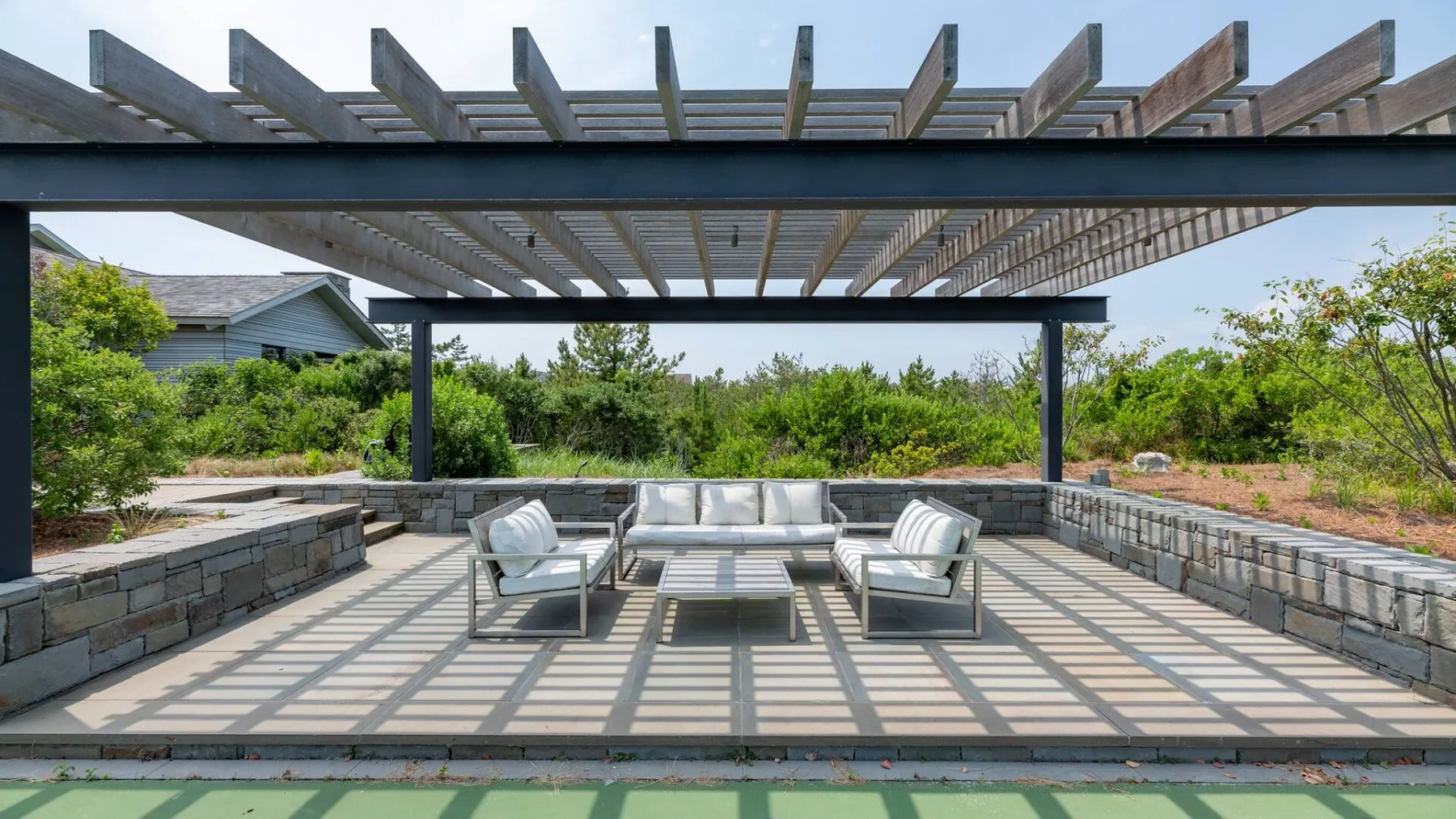 Una zona de estar exterior a la sombra de una pérgola, con una mesa baja y vegetación alrededor.