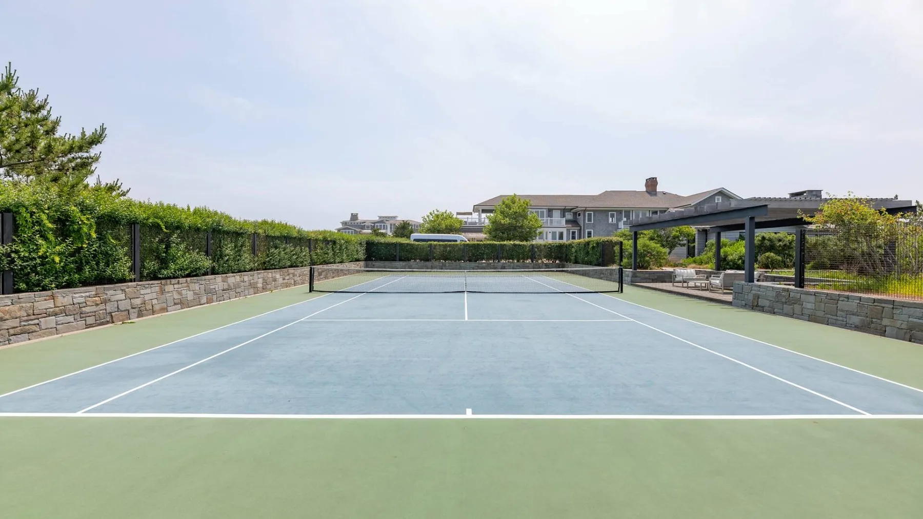 An outdoor tennis court is bordered by hedges and low stone walls.