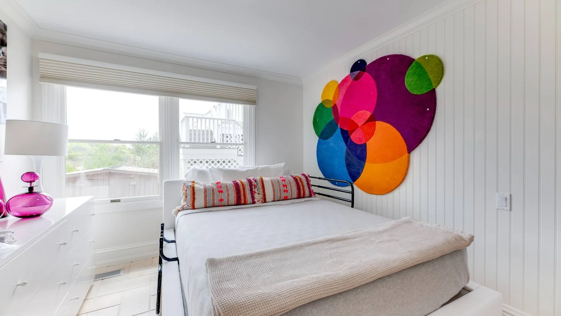 Un dormitorio luminoso tiene una cama bien tendida junto a una gran decoración colorida en la pared.