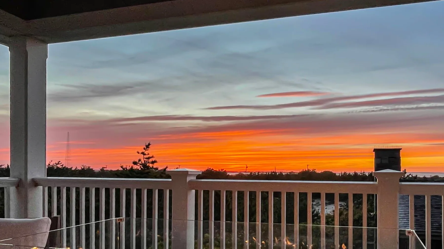 Un balcón cubierto da a una barandilla y árboles bajo un cielo de atardecer.