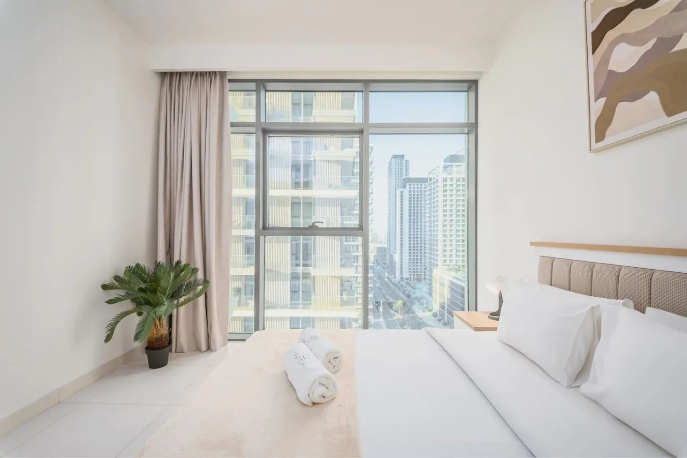 Un dormitorio luminoso con una cama junto a una gran ventana con vistas a edificios de la ciudad.