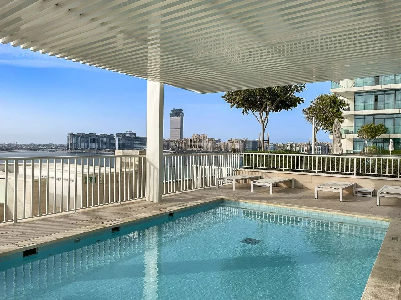 Una piscina está bajo una terraza sombreada con vistas a los edificios cercanos.