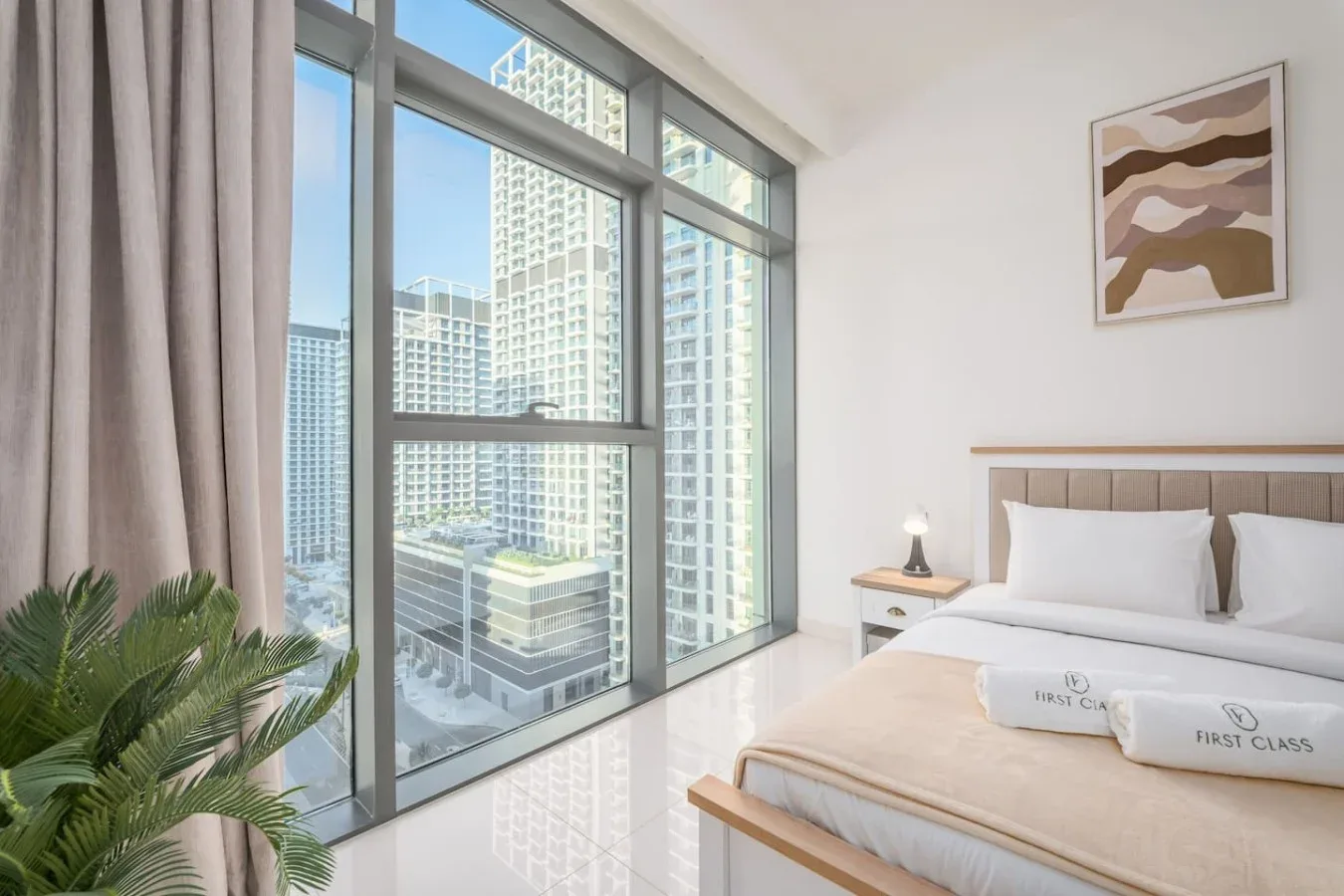 Un dormitorio luminoso con una cama junto a grandes ventanales con vistas a edificios de la ciudad.