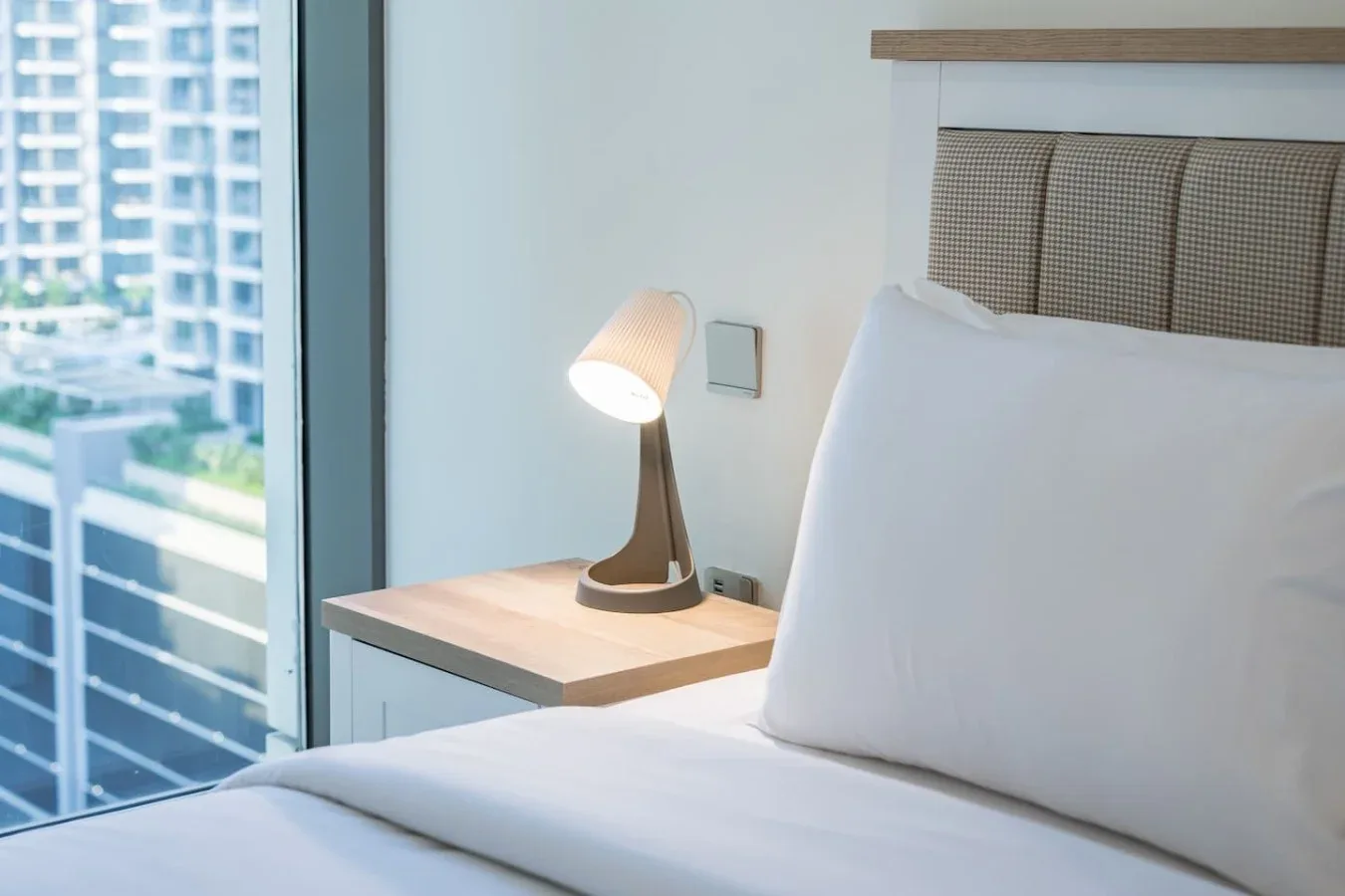 Un dormitorio con ropa de cama blanca, una lámpara de noche y una ventana grande.