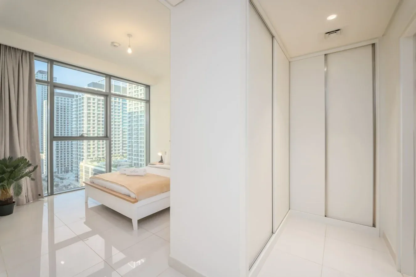 Un dormitorio luminoso con una cama junto a grandes ventanas con vistas a edificios cercanos.