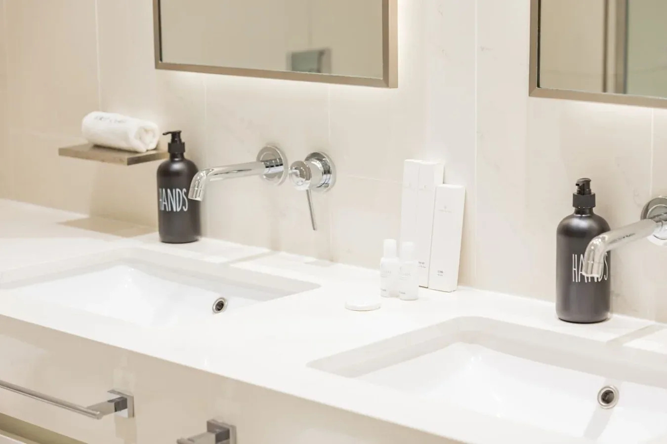 Un tocador de baño con dos lavabos, grifos de pared y dispensadores oscuros de jabón.