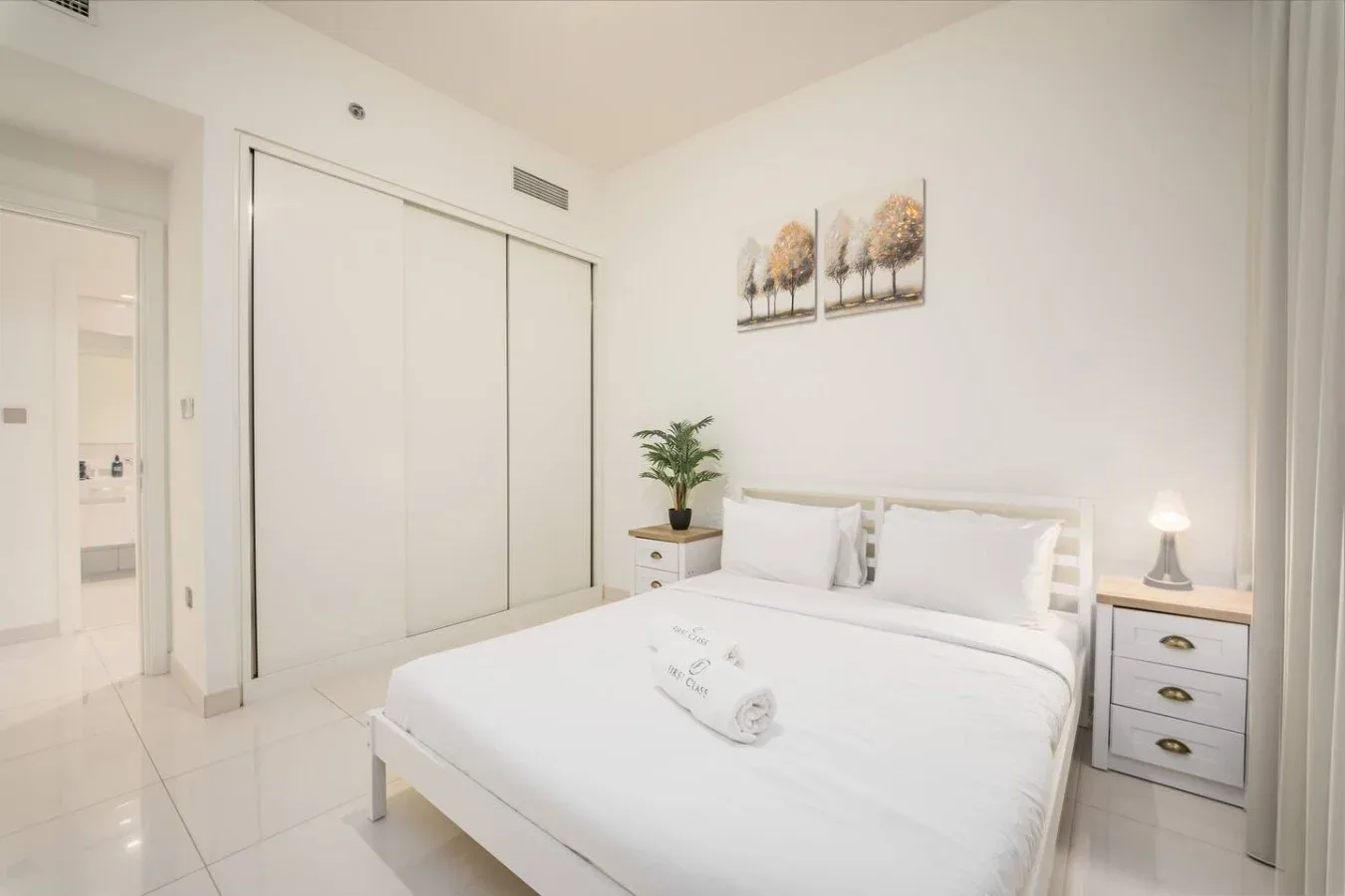 Un dormitorio luminoso con una cama blanca, puertas correderas de armario y una mesita de noche.