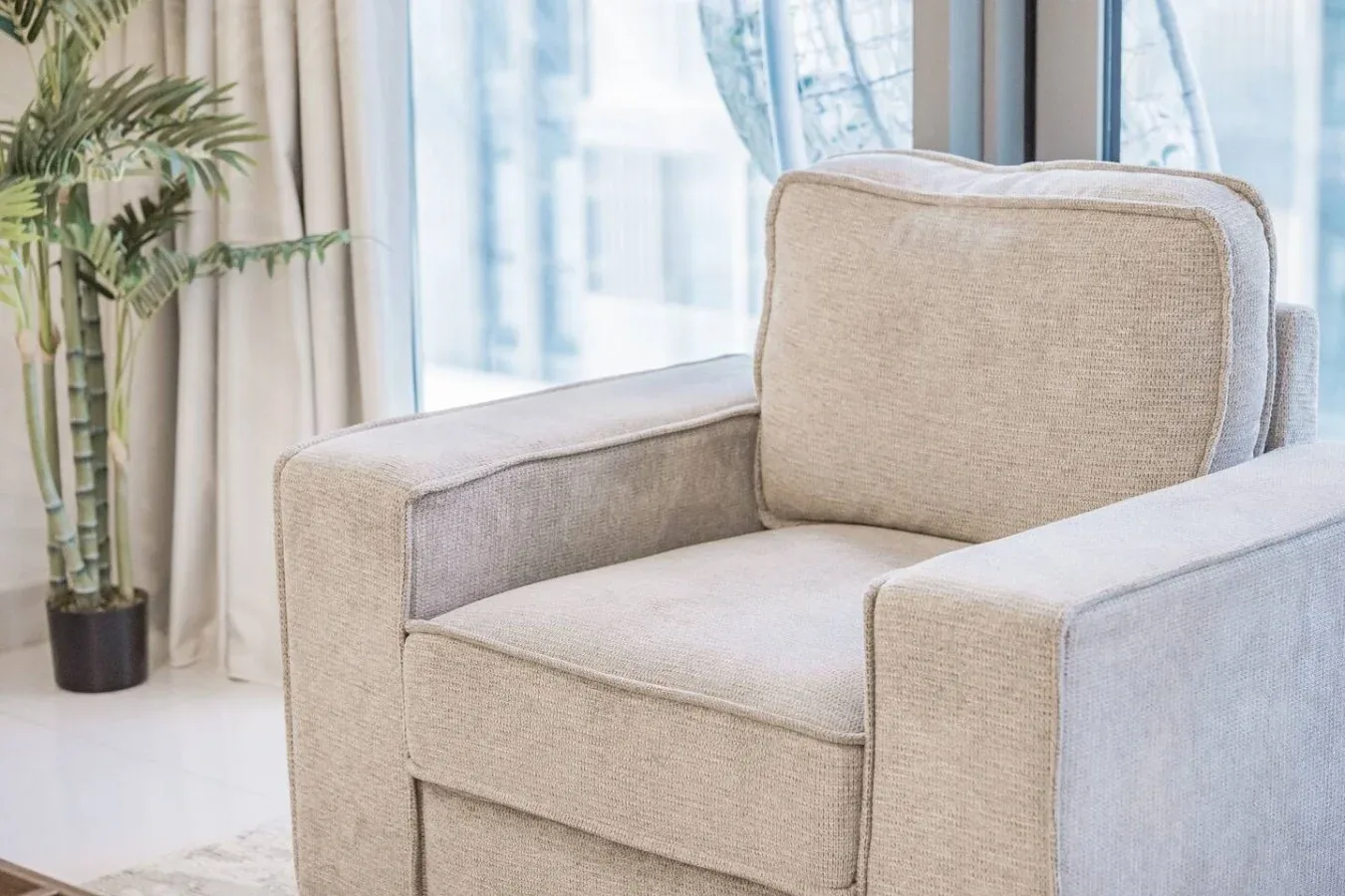Un sillón tapizado claro está junto a una ventana grande y una planta en maceta.