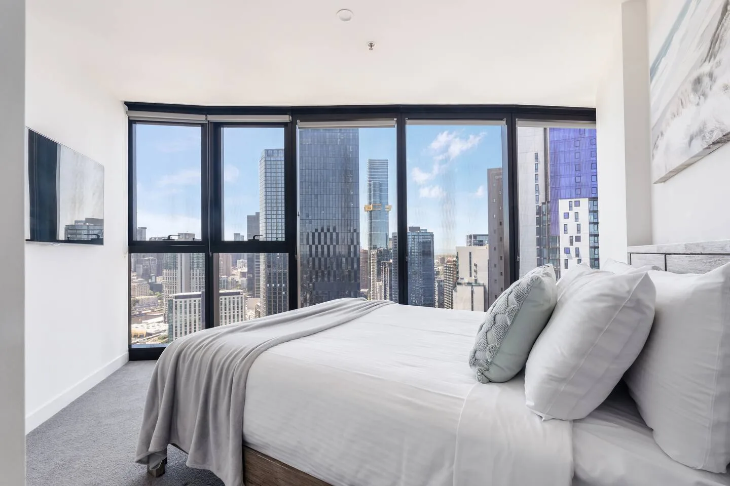 Un dormitorio luminoso con una cama junto a grandes ventanas con vistas a edificios de la ciudad.