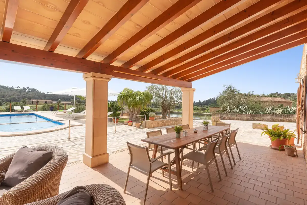 A covered patio with a dining table overlooks a swimming pool and garden.