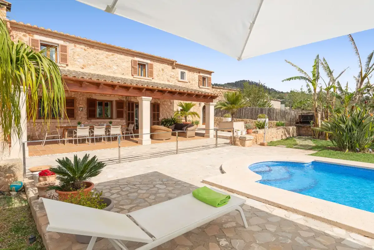 A sunny patio with lounge seating overlooks a swimming pool and stone house.