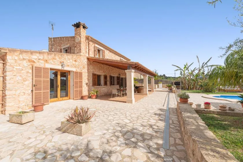 A stone house opens onto a paved patio with a covered seating area and pool.