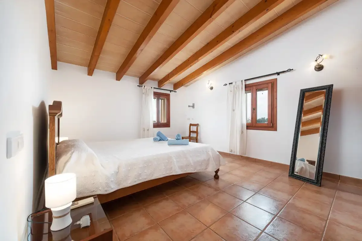 A bright bedroom with a bed, exposed wooden ceiling beams, and tiled flooring.