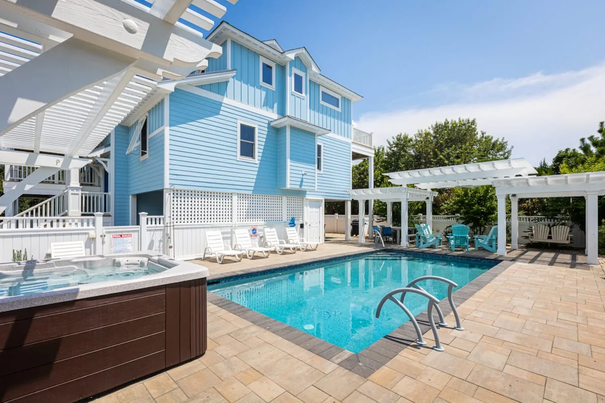 Una piscina al aire libre junto a una casa azul, con tumbonas y una pérgola.