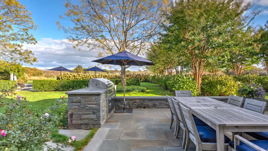 Un patio exterior con mesa de comedor, sombrilla y barbacoa de piedra junto a un jardín.