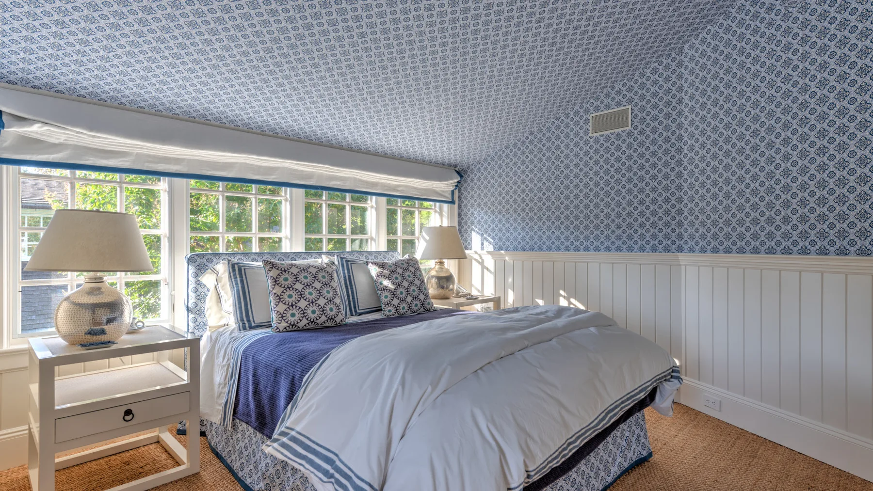 Un dormitorio con una cama bajo grandes ventanas y papel tapiz estampado.