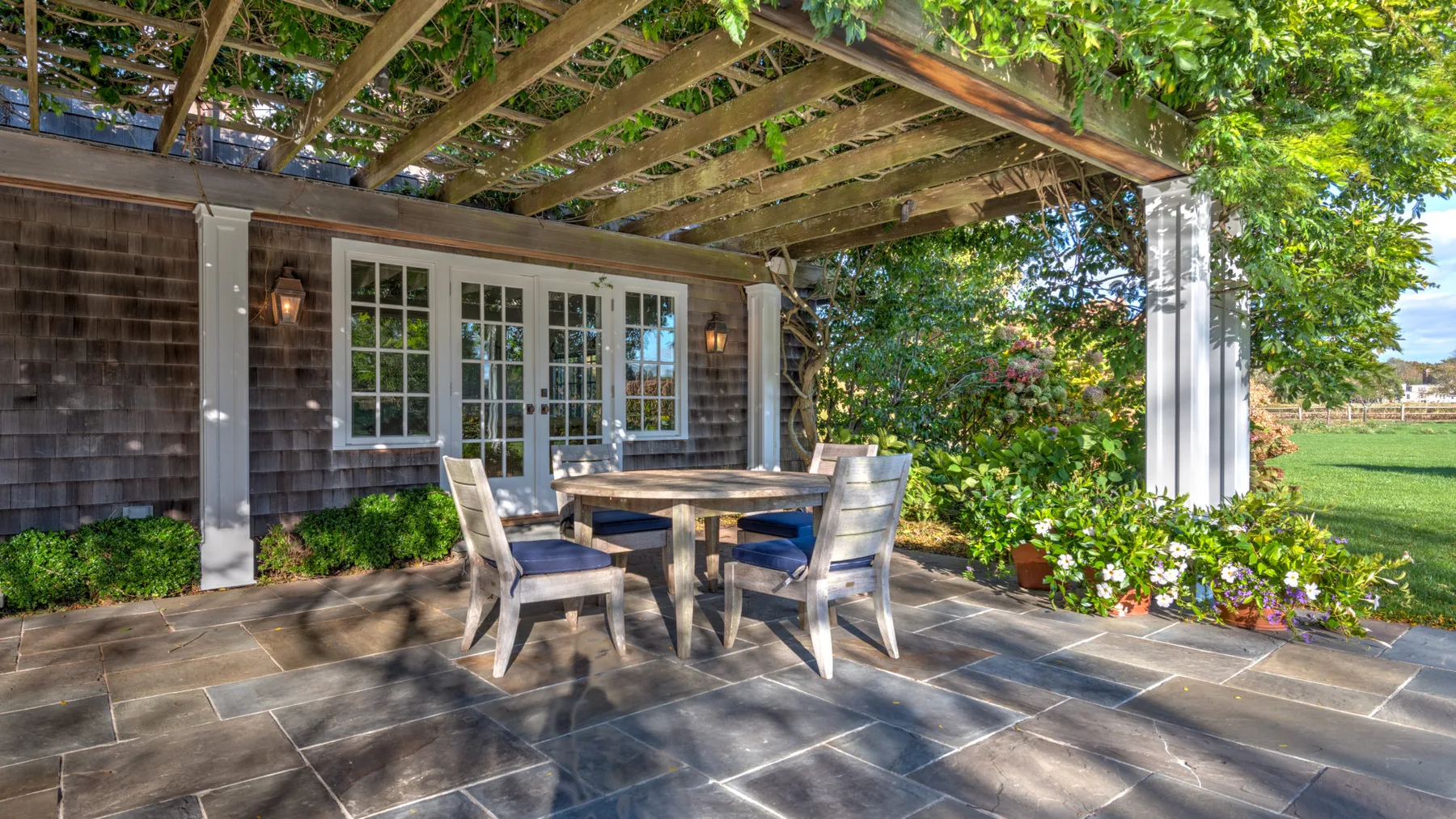 Un patio exterior cubierto tiene una mesa de comedor y asientos junto a la casa.