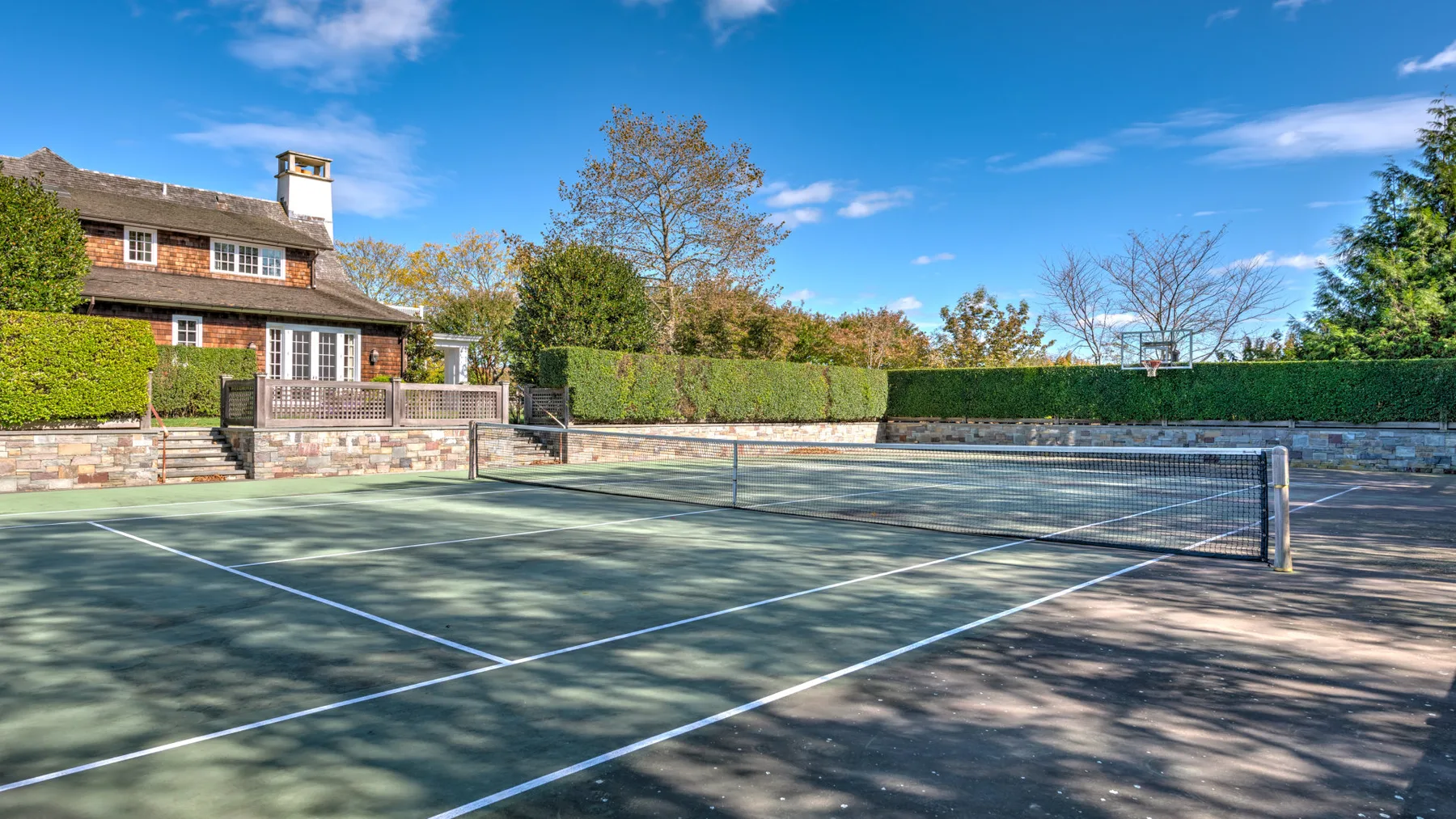 Una pista de tenis al aire libre está junto a una casa de ladrillo y setos recortados.