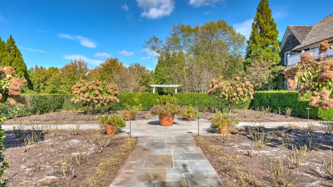 Un sendero de jardín atraviesa parterres y conduce hacia un seto y una casa.
