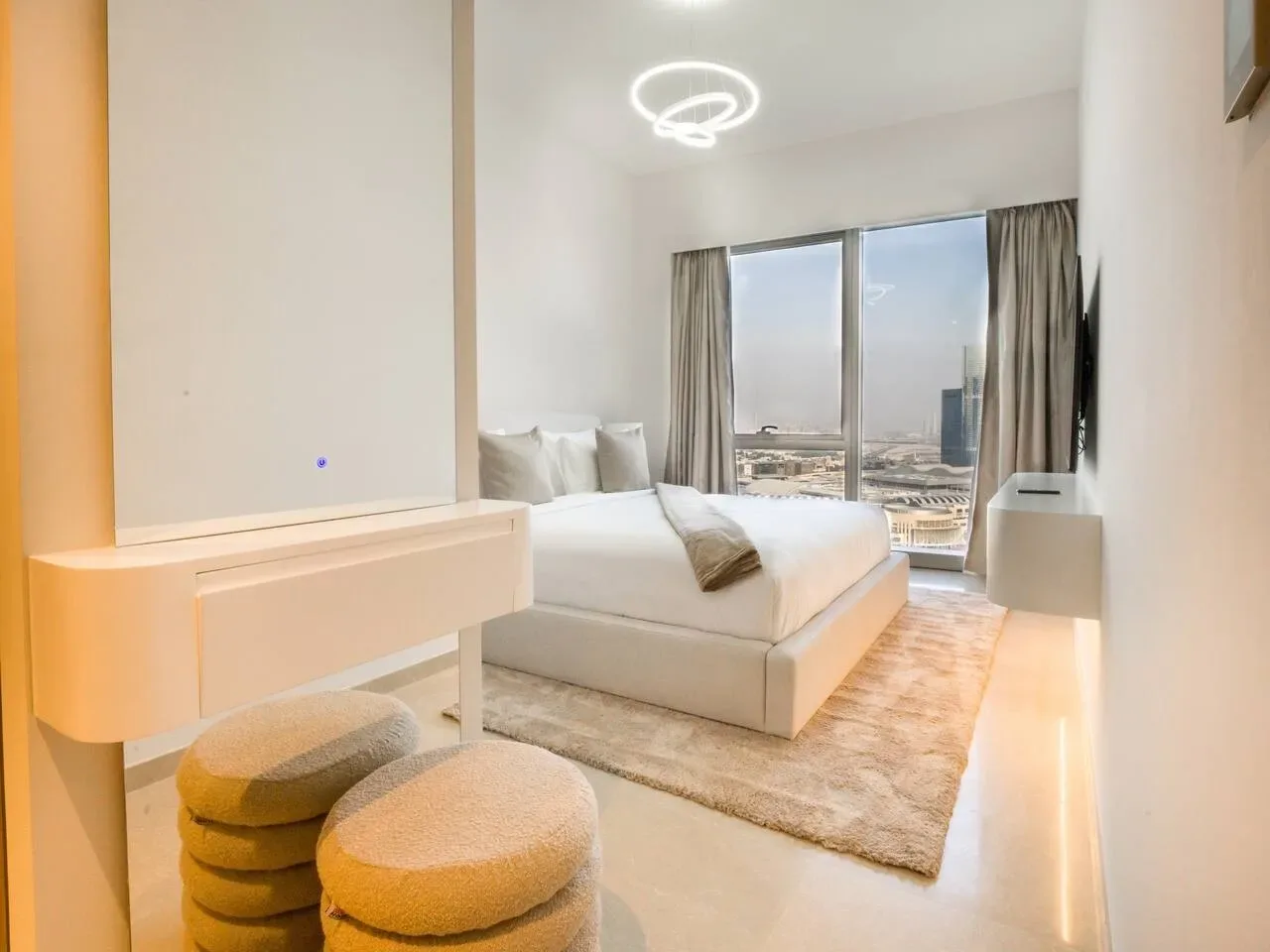 A bright bedroom with a bed beside large windows overlooking the city.