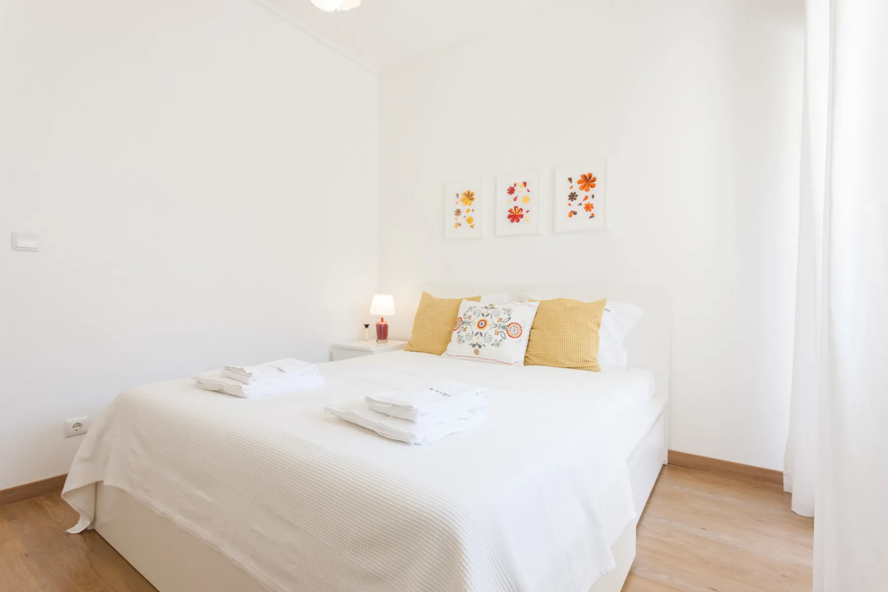 Un dormitorio luminoso con una cama bien tendida y cuadros coloridos encima.