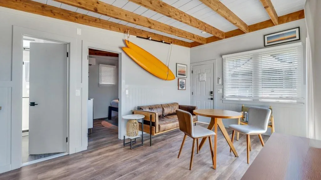A bright dining and sitting area has a wooden table, seating, and exposed ceiling beams.