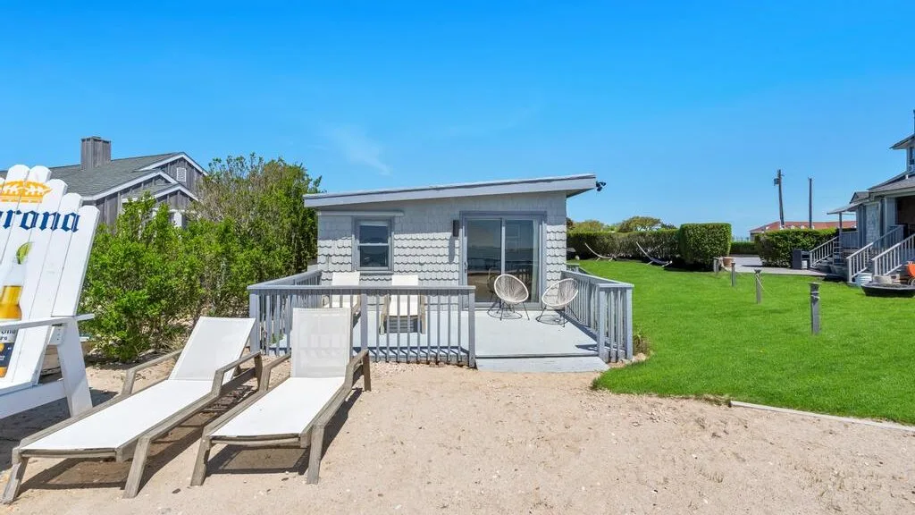 A small building opens onto a sandy patio with lounge chairs and a grassy lawn.