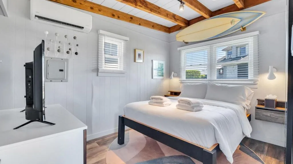 A bright bedroom with a neatly made bed and a surfboard mounted above the windows.