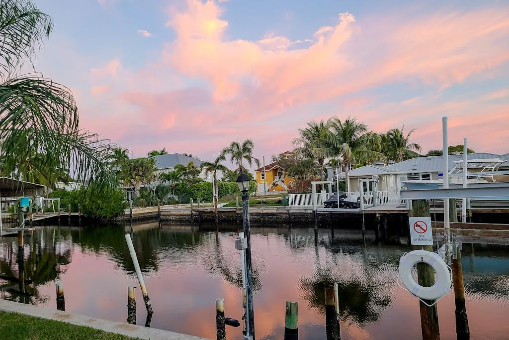 A waterfront canal with boat docks and palm trees beneath a pink sunset sky.