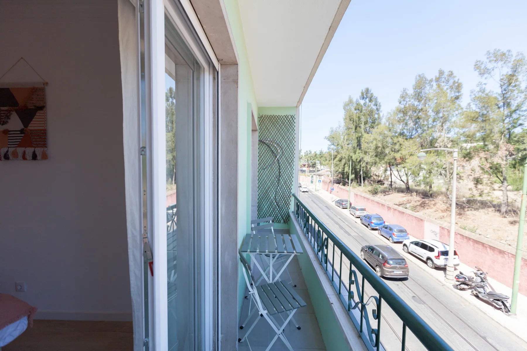 A narrow balcony with folding seating overlooks a tree-lined street.