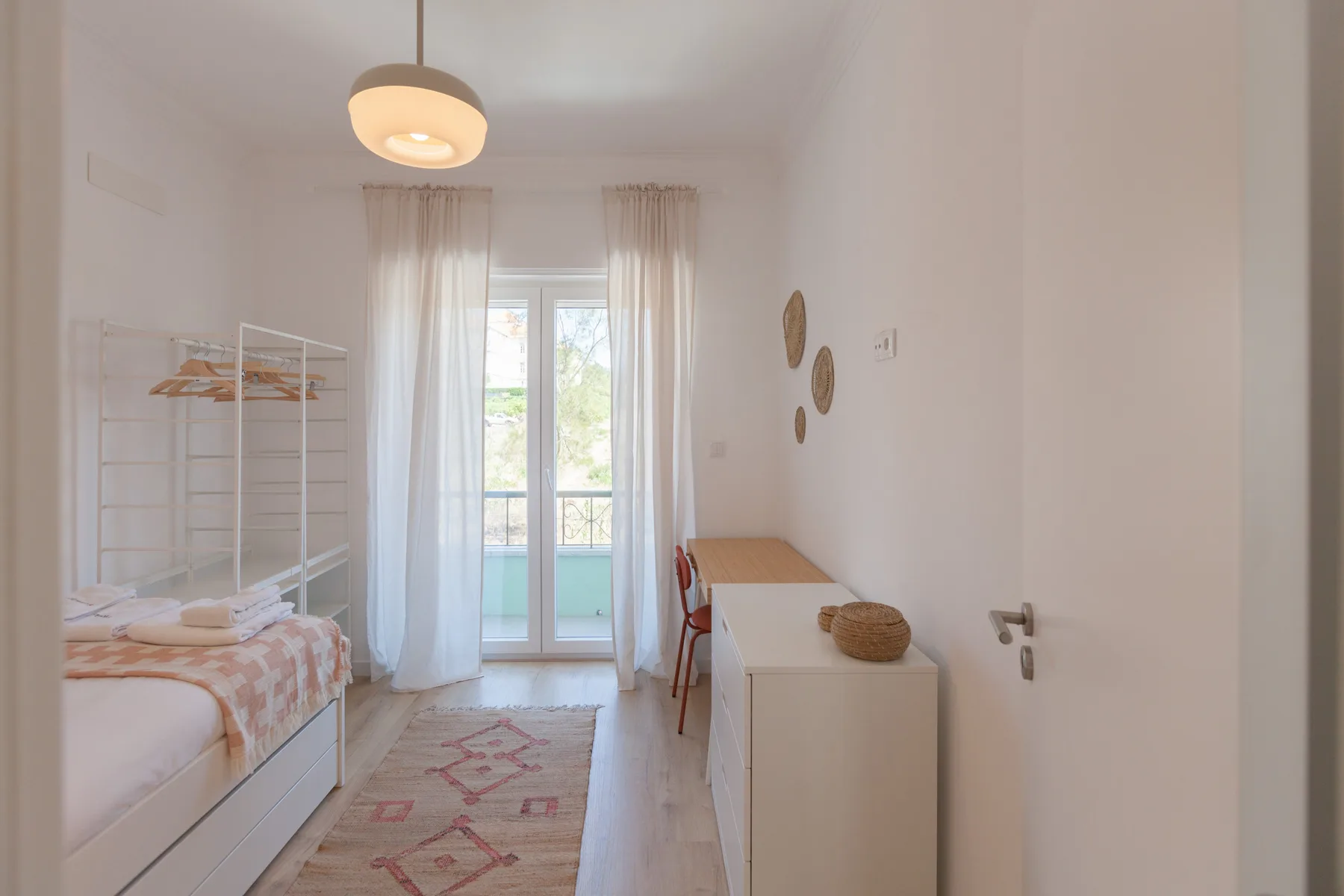 A bright bedroom with a bed, desk, and glass doors opening to a balcony.