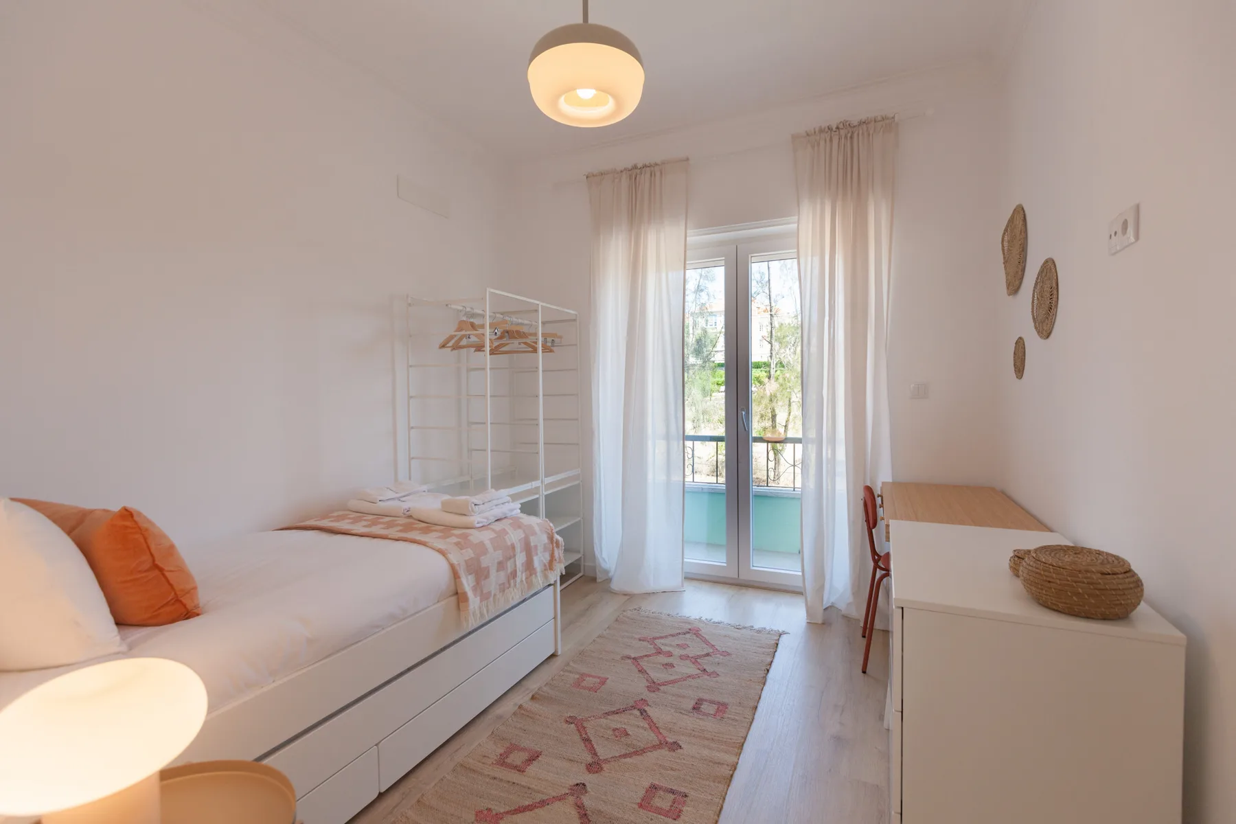 A bright bedroom with a bed, a desk, and glass doors opening onto a balcony.