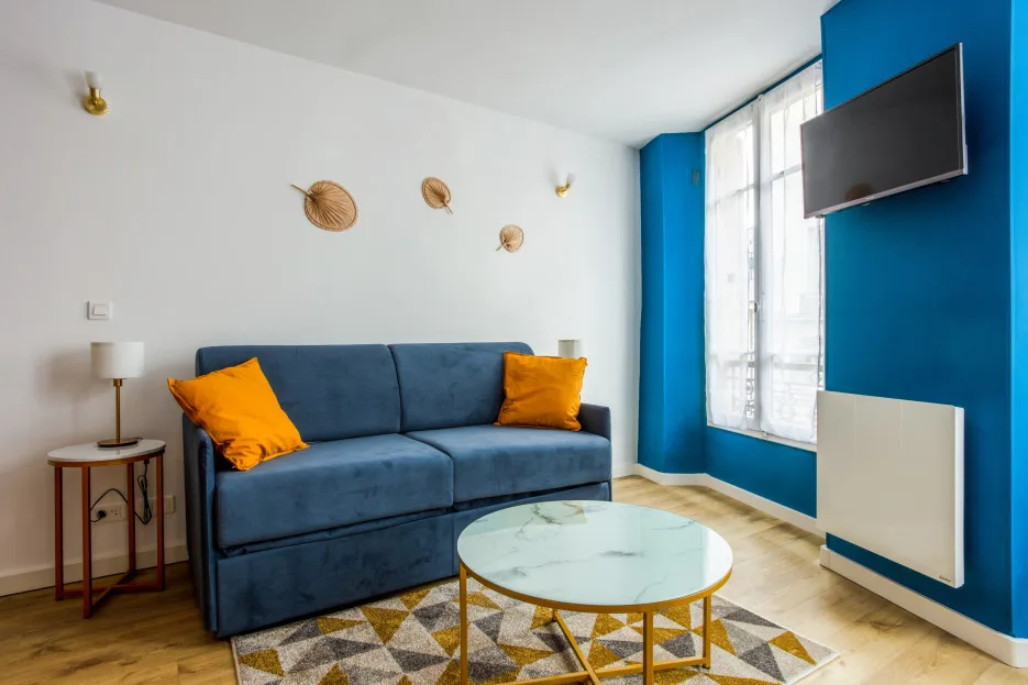 Una sala de estar con asientos azules, una mesa de centro redonda y un televisor en la pared.