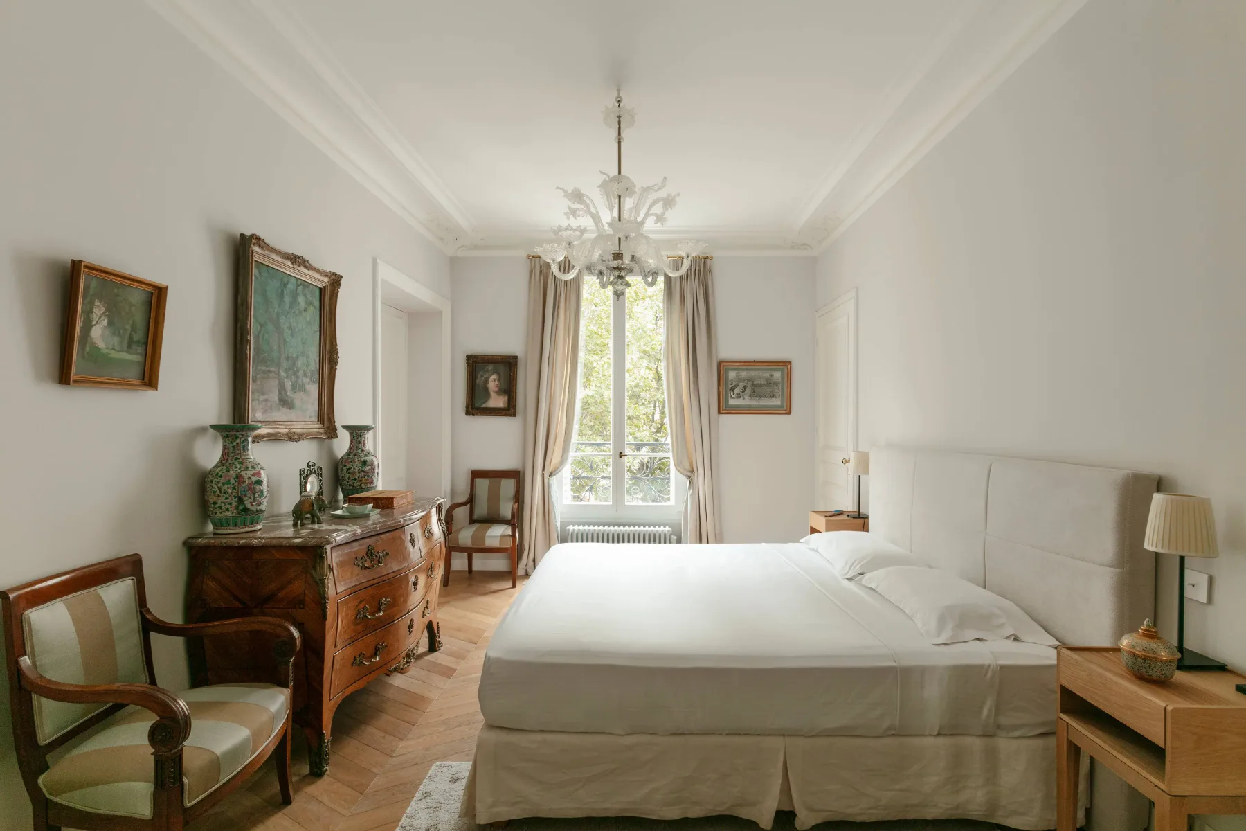 A bright bedroom with a large bed, tall window, and antique-style furnishings.