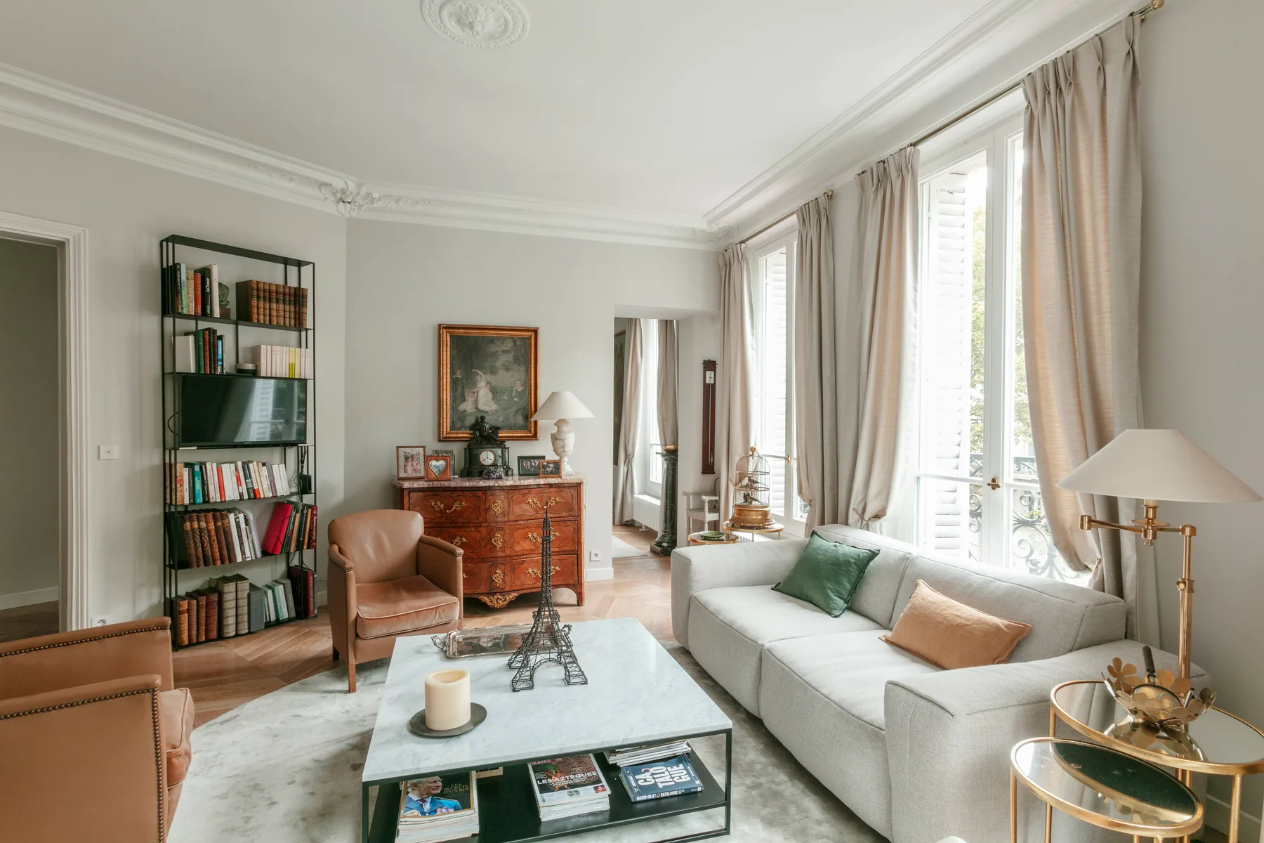 A bright living room with seating, a bookcase, and tall windows.