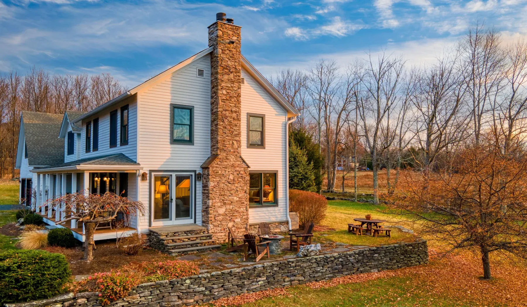 Una casa blanca con una chimenea de piedra se alza junto a un patio con una mesa de picnic.