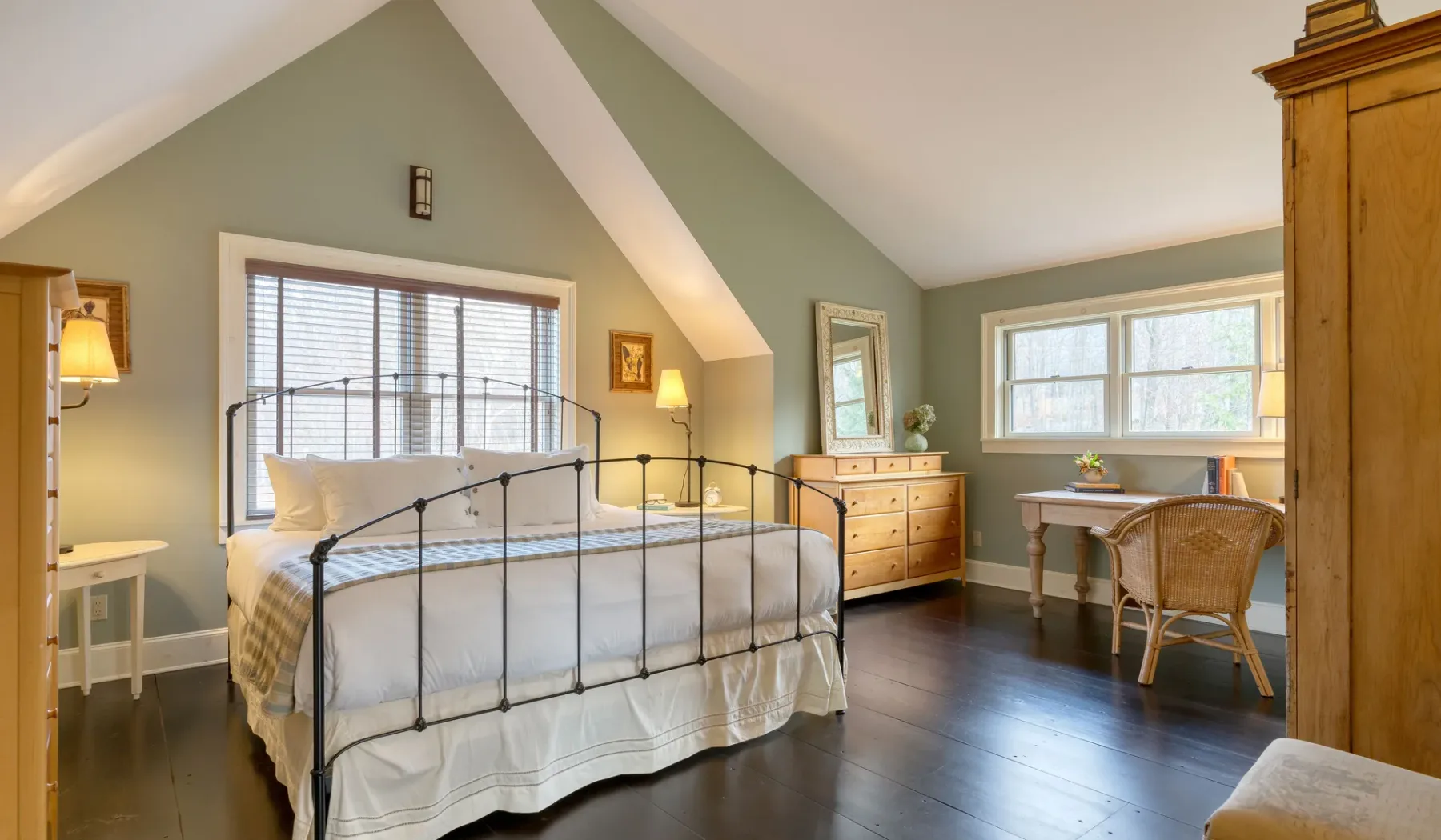 Un dormitorio con una cama de metal, una cómoda y una mesa pequeña junto a ventanas luminosas.