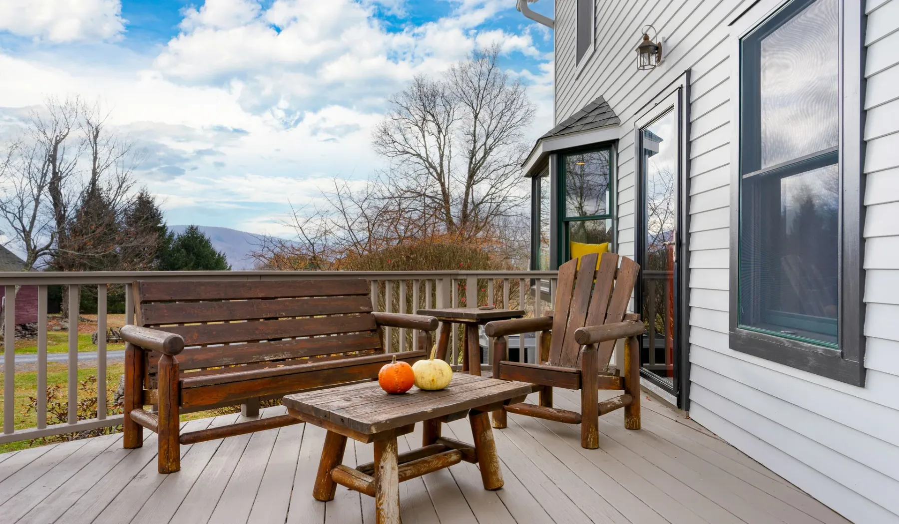 Una terraza de madera junto a una casa tiene un banco, sillas y una mesa pequeña.