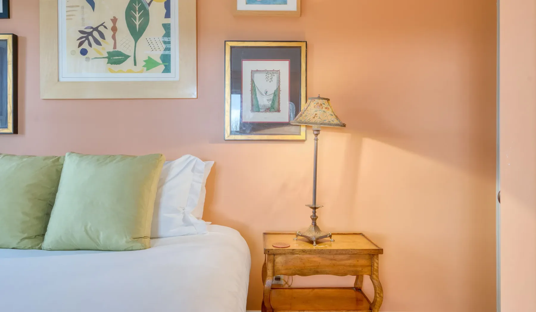 Un dormitorio con una cama tendida, cuadros enmarcados y una mesita con una lámpara.