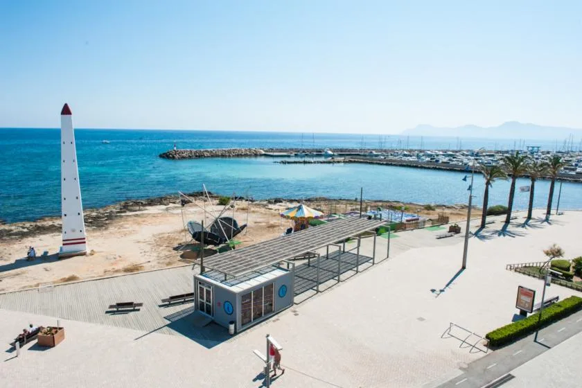 Un paseo junto al mar mira hacia aguas turquesas, con una zona cubierta para sentarse y palmeras.